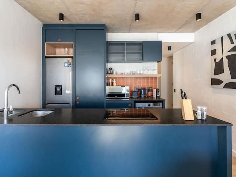Modern kitchen featuring dark blue cabinetry, a central island with a hob, stainless steel sink, and a stylish tiled backsplash under an exposed concrete ceiling.
