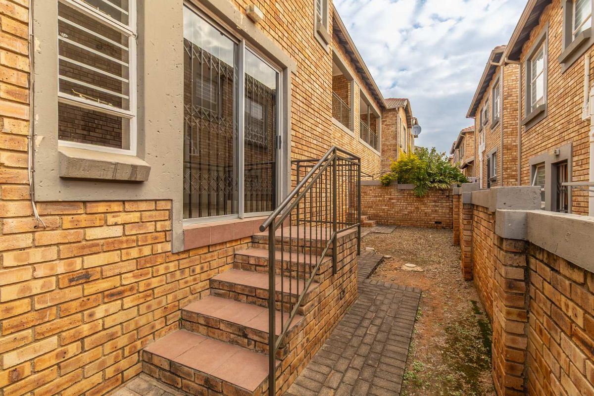 A rear exterior view of the townhouse featuring a brick facade, paved walkway, and a small staircase leading to a sliding glass door entrance.