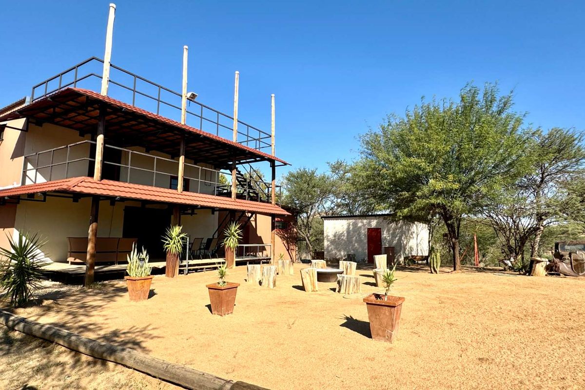 A spacious rear exterior view featuring a two-story building with a wrap-around balcony, covered patio area, and a large, open sandy garden space.