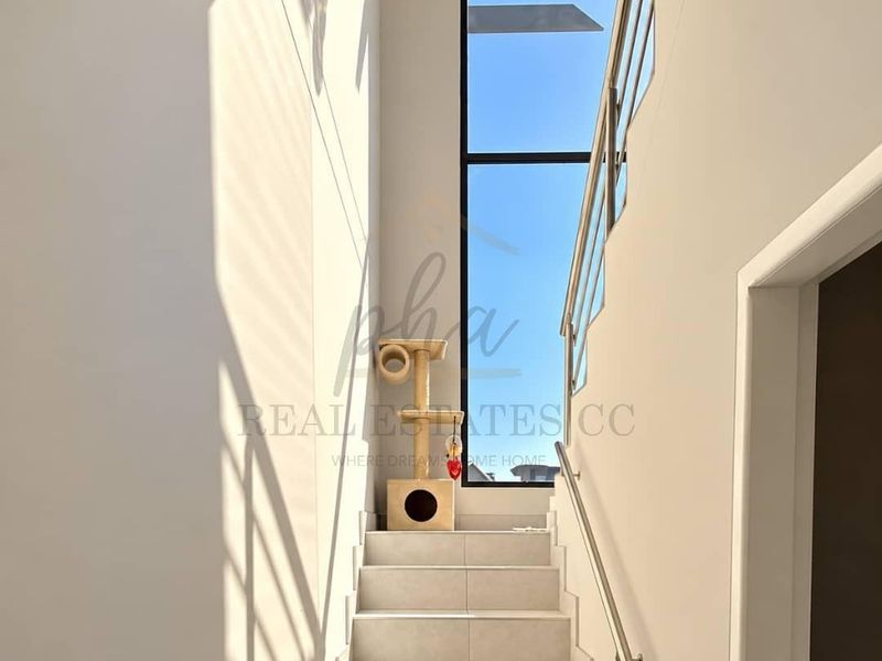 A bright, modern staircase featuring tiled steps, a sleek metal handrail, and a large full-height window that allows for abundant natural light.