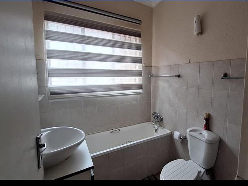 A functional bathroom featuring a built-in bathtub, wall-mounted basin, toilet, and tiled wall finishes with a roller blind for privacy.