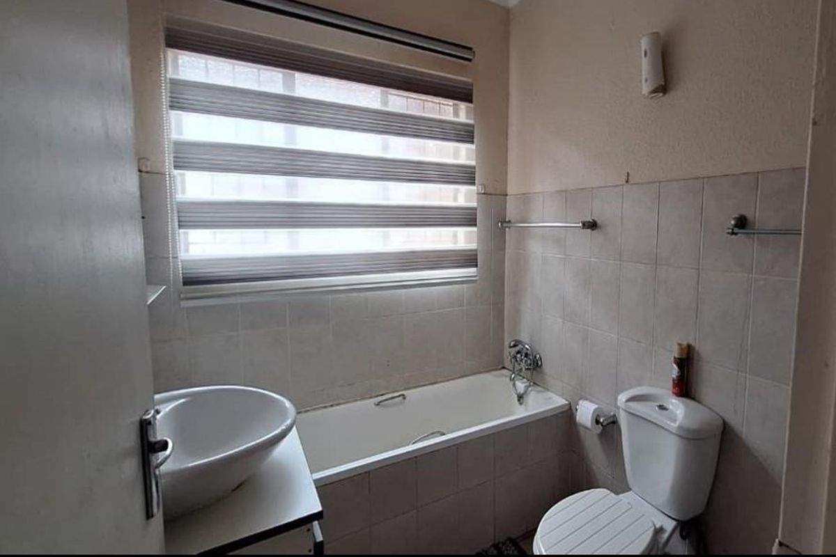 A functional bathroom featuring a built-in bathtub, wall-mounted basin, toilet, and tiled wall finishes with a roller blind for privacy.