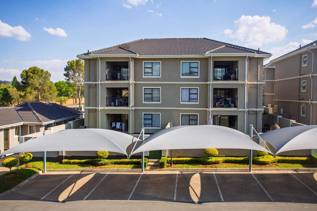 Exterior view of a multi-story apartment building featuring covered parking bays, landscaped garden beds, and private balconies for each unit.