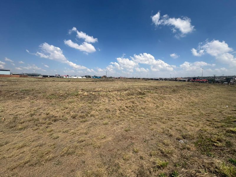 A wide-angle view of a vacant commercial land plot featuring open grass terrain with surrounding industrial and commercial infrastructure in the distance.