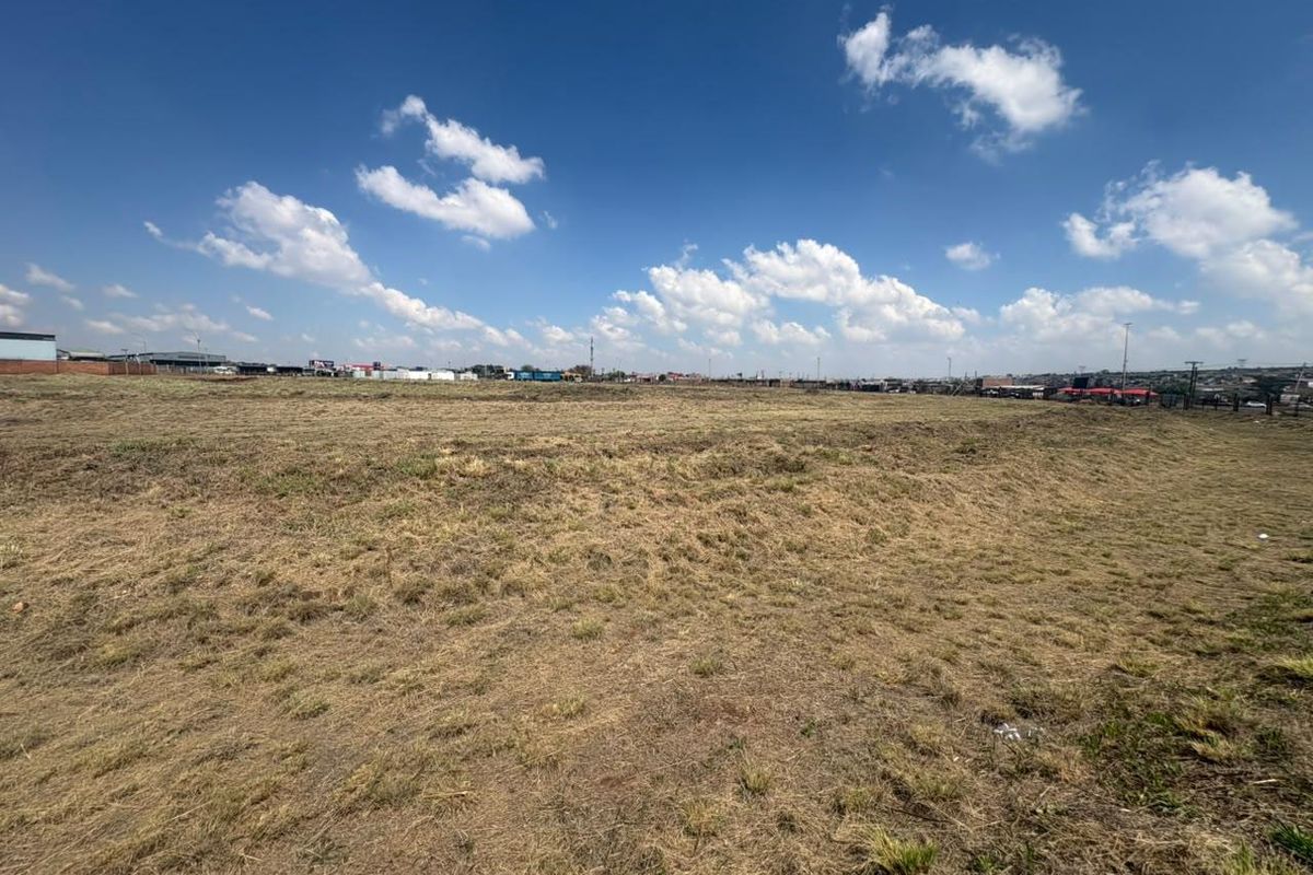 A wide-angle view of a vacant commercial land plot featuring open grass terrain with surrounding industrial and commercial infrastructure in the distance.