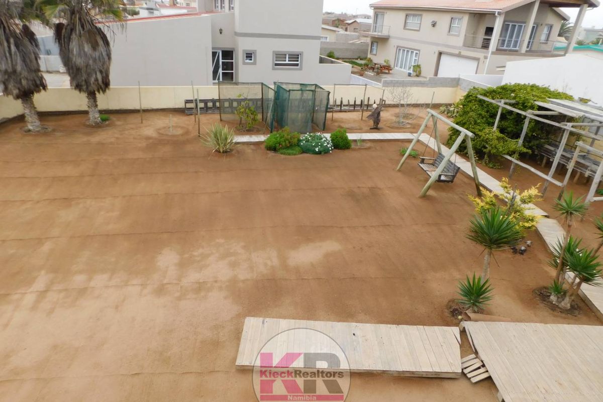 An elevated view of a large, unlandscaped rear garden area featuring a wooden deck, perimeter wall, and surrounding residential properties.