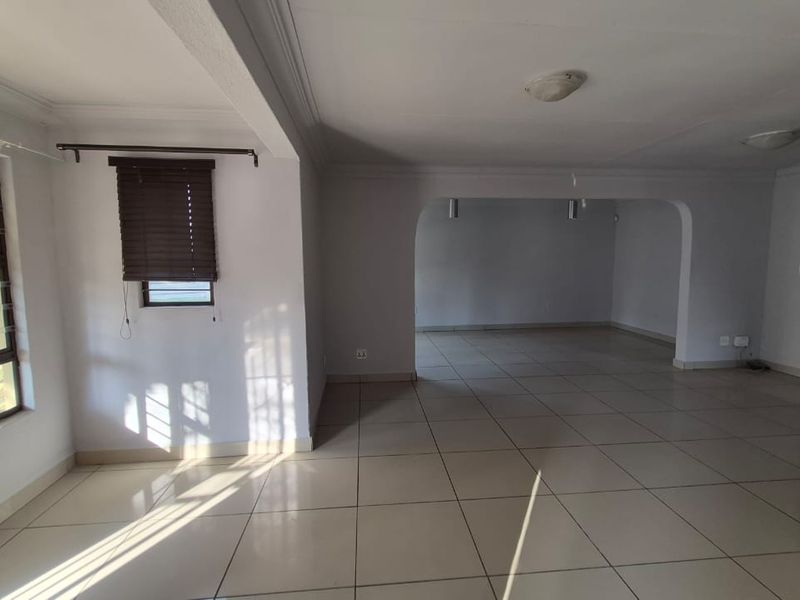 A spacious, empty living area featuring neutral tiled flooring, an open-plan archway design, and natural light through large windows with security bars.