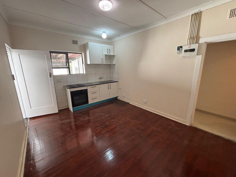 A compact, empty kitchen featuring timber flooring, built-in cabinetry with an integrated oven and hob, and a tiled splashback.