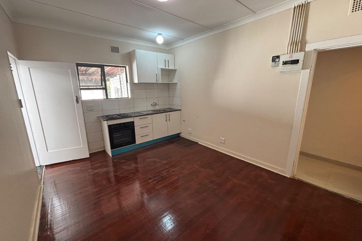 A compact, empty kitchen featuring timber flooring, built-in cabinetry with an integrated oven and hob, and a tiled splashback.