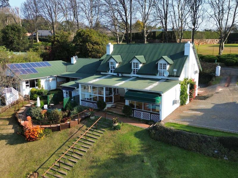 Aerial view of a well-maintained property featuring a green corrugated metal roof, solar panels, dormer windows, and an expansive landscaped garden with paved driveway.