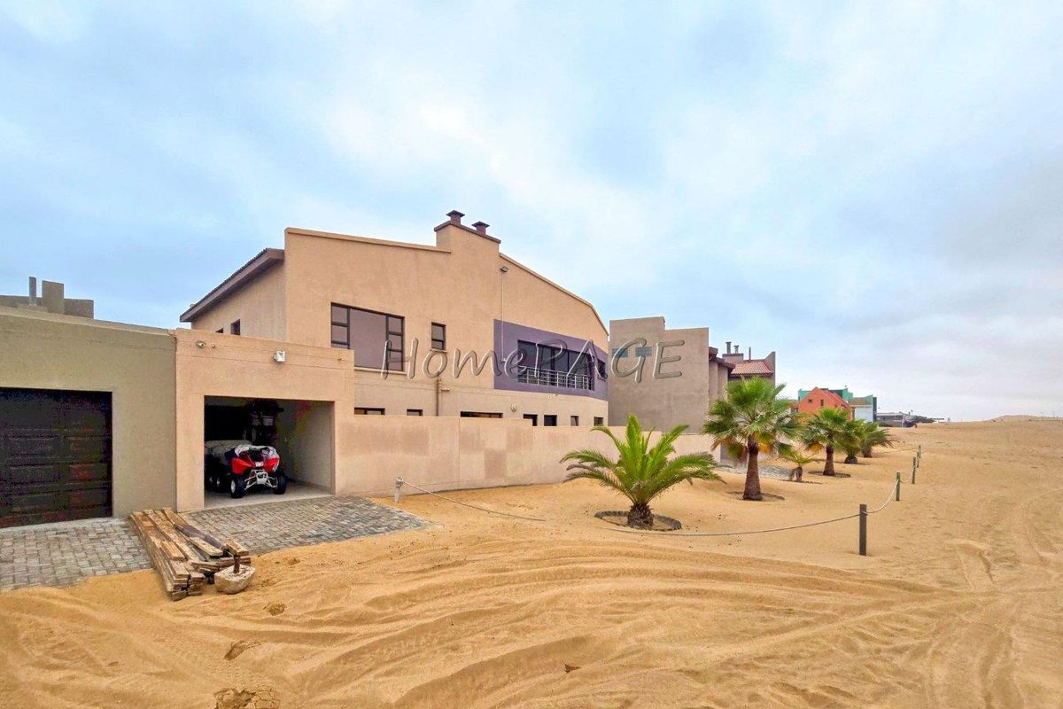Exterior view of a modern multi-story house featuring a garage, sandy surroundings, and landscaped palm trees.