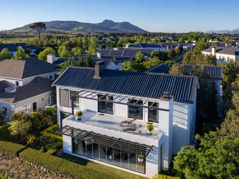 Aerial view of a modern two-story house featuring a metal roof, glass balustrades, and a spacious upper-level balcony overlooking a landscaped residential estate.