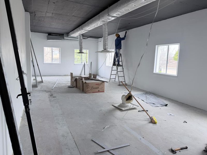 An unfinished office space featuring exposed ceiling ductwork, concrete flooring, and large windows, currently undergoing renovation and painting.