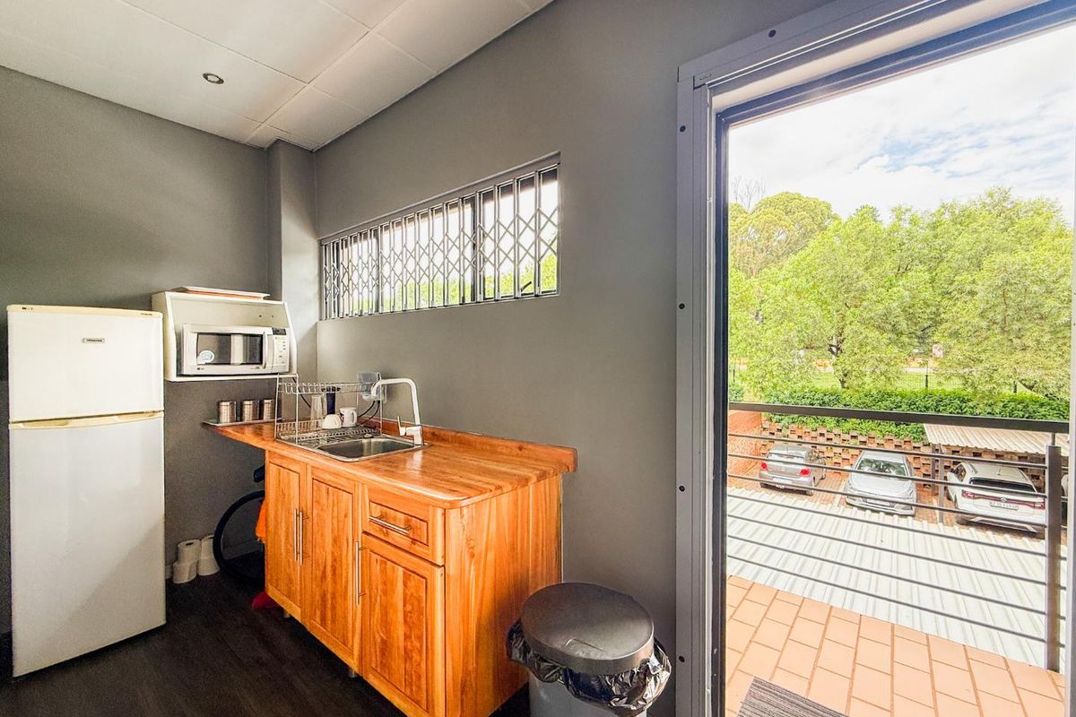 A compact office kitchenette featuring wooden cabinetry, a stainless steel sink, and a window with security bars, opening onto a balcony.