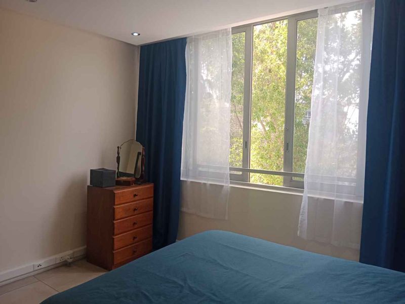 A bedroom featuring tiled flooring, recessed downlights, and large aluminium-framed windows providing natural light and a view of the surrounding trees.