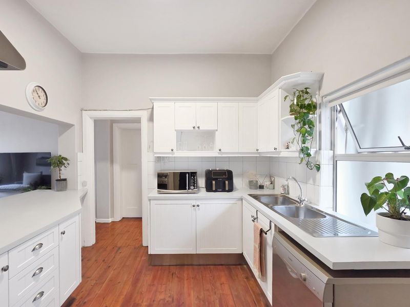 A bright, functional kitchen featuring white cabinetry, timber flooring, and a stainless steel double sink with a tiled backsplash.