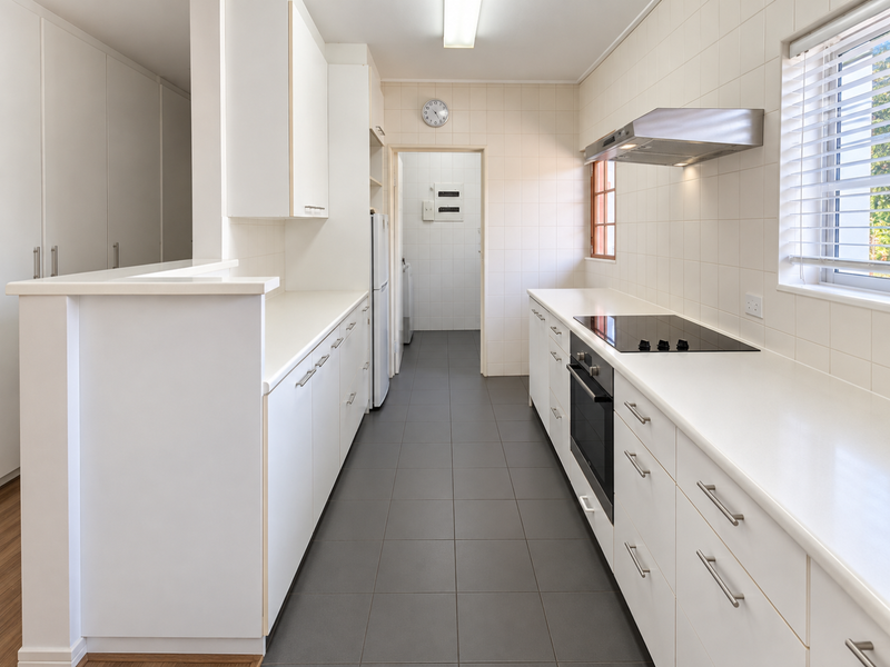 A clean, functional kitchen featuring white cabinetry, stone countertops, a built-in oven, electric hob, and tiled flooring, leading to a separate utility area.