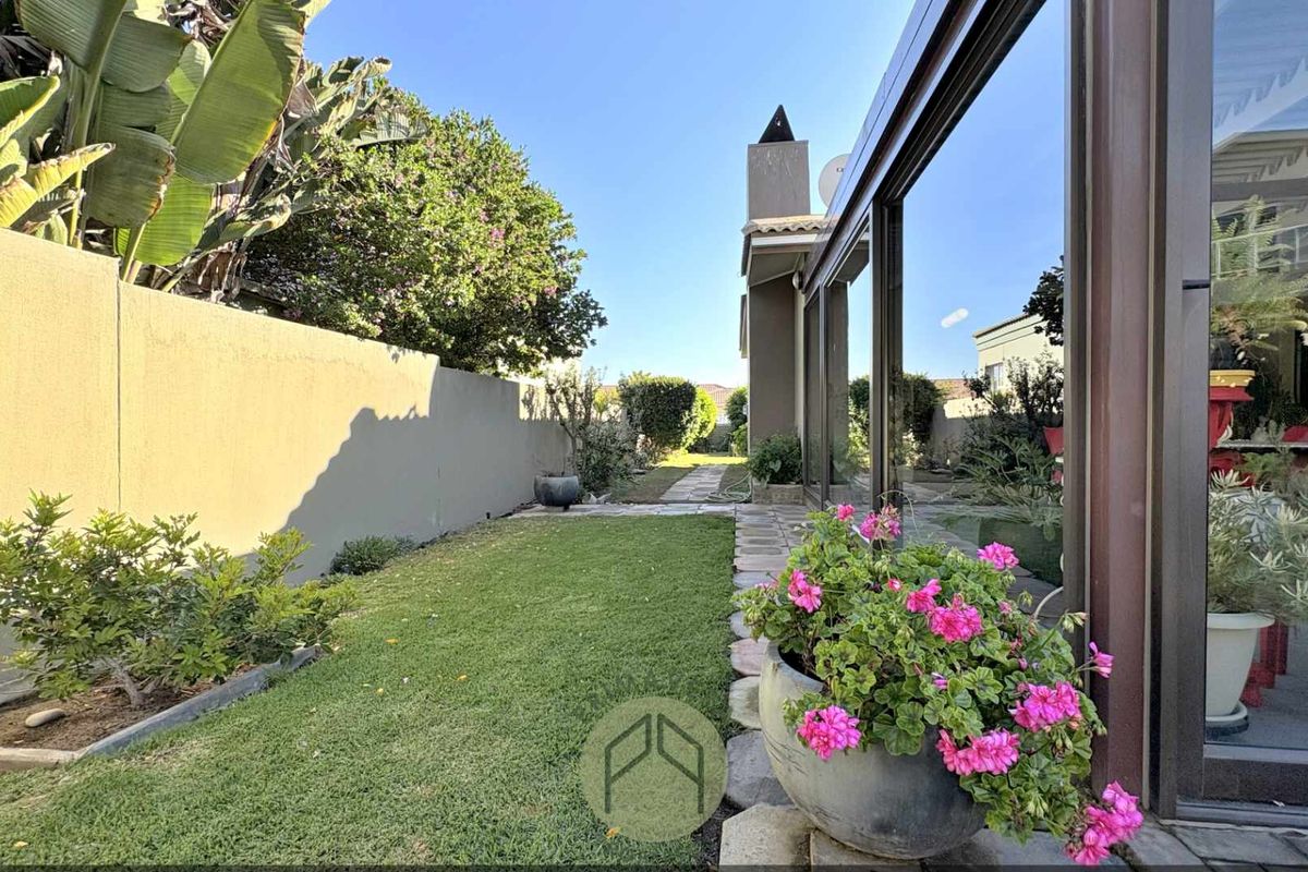 A well-maintained rear garden featuring a paved walkway, boundary wall, and an outdoor fireplace, adjacent to large sliding glass doors.