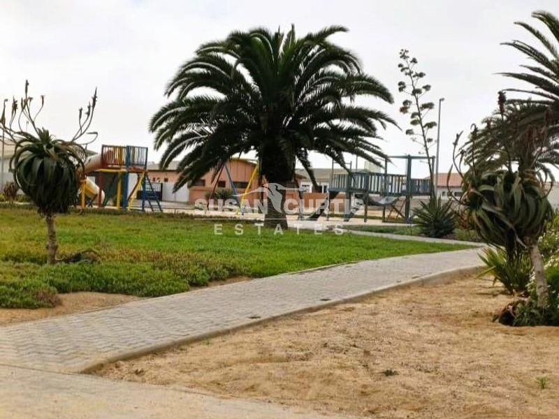 A landscaped garden area featuring a paved walkway, mature palm trees, and a children's play area in the background.
