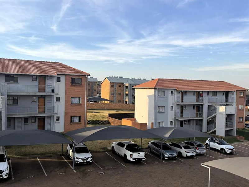 Exterior view of a multi-story apartment complex featuring brick and plaster facades, tiled roofs, and dedicated paved parking bays with shade cloth structures.