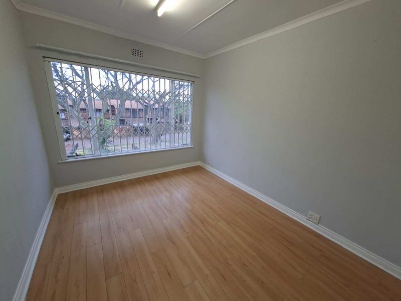 A bright, empty bedroom featuring laminate flooring, neutral walls, and a large window fitted with a security gate.