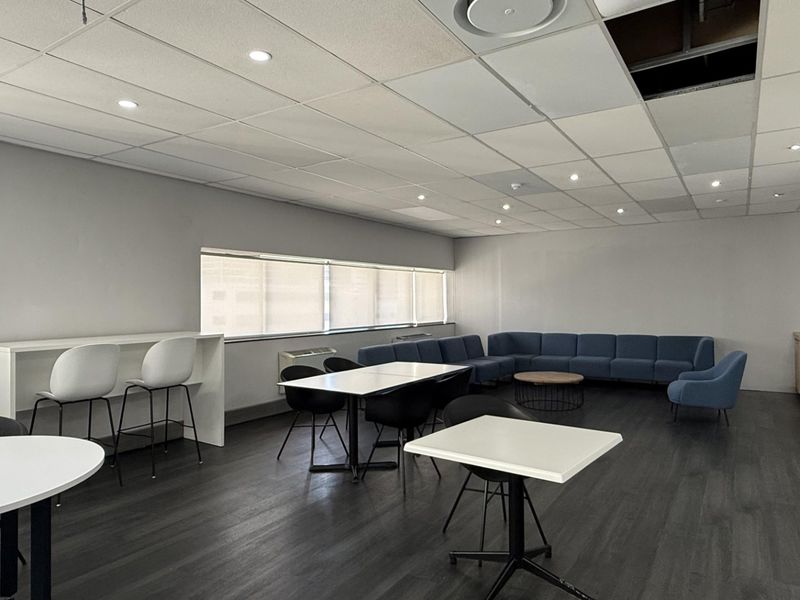 A spacious open-plan office area featuring dark vinyl flooring, recessed lighting, and a suspended ceiling with a missing tile.