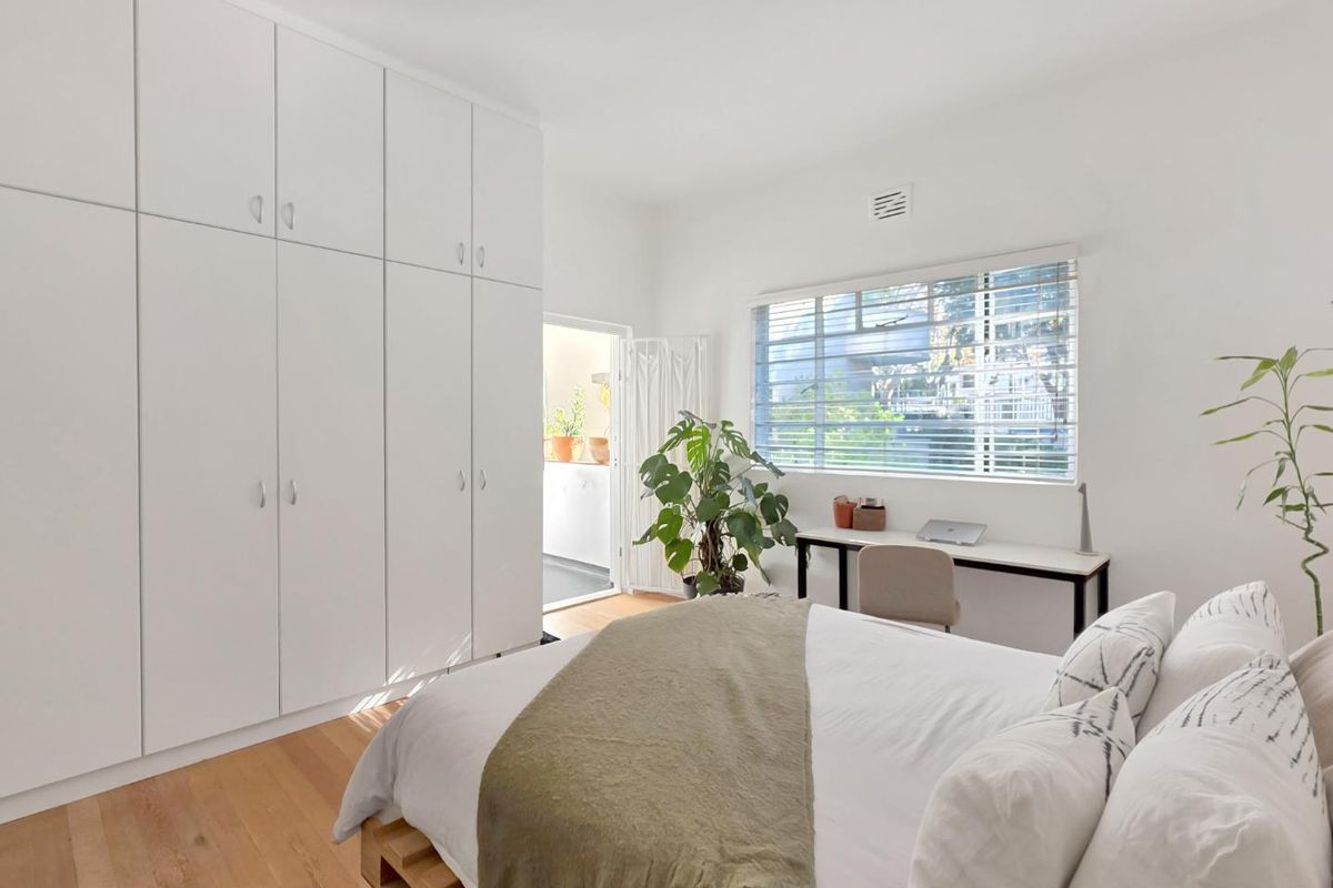 Bright bedroom featuring extensive built-in floor-to-ceiling wardrobes, timber flooring, and large windows with venetian blinds, offering a clean and modern aesthetic.