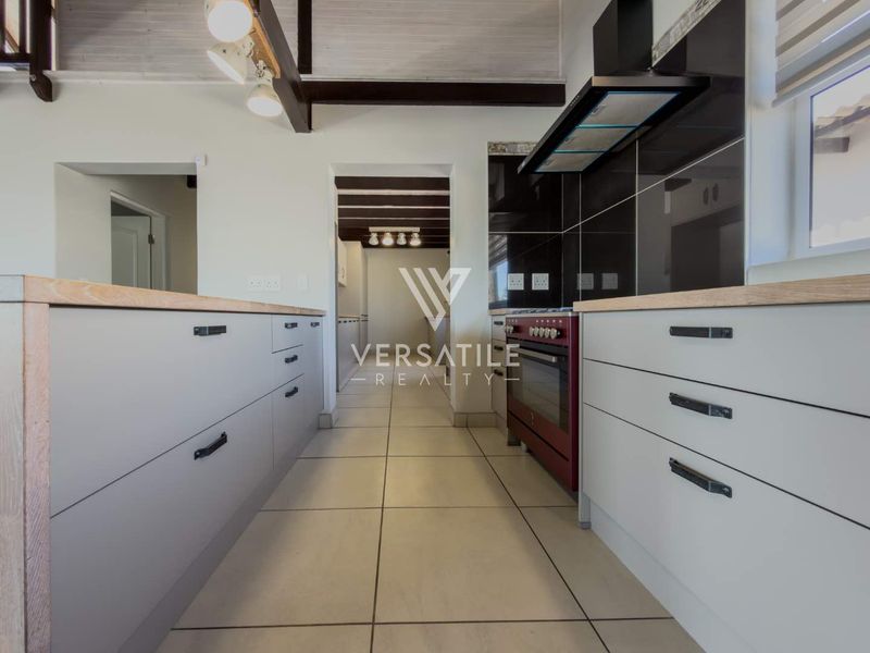 Modern kitchen featuring light grey cabinetry, timber countertops, tiled flooring, and exposed ceiling beams, leading into a secondary utility or scullery area.