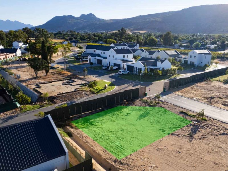 An aerial view of a vacant residential plot in a developed estate, featuring road access and surrounding mountain views.