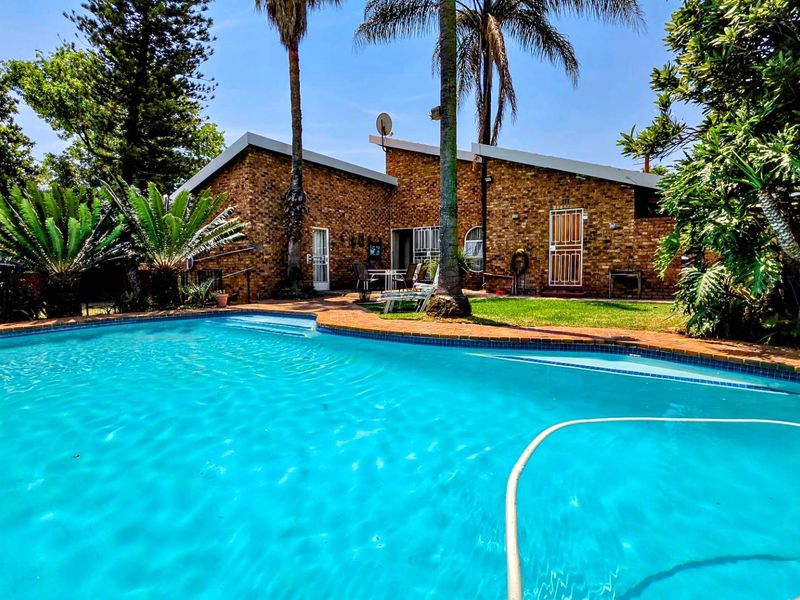 A well-maintained swimming pool area featuring a brick exterior facade, mature landscaping, and a clear view of the rear of the house.