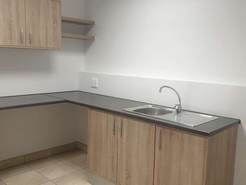 A clean, functional kitchenette area featuring light wood-tone cabinetry, dark laminate countertops, a stainless steel sink, and neutral tiled flooring.