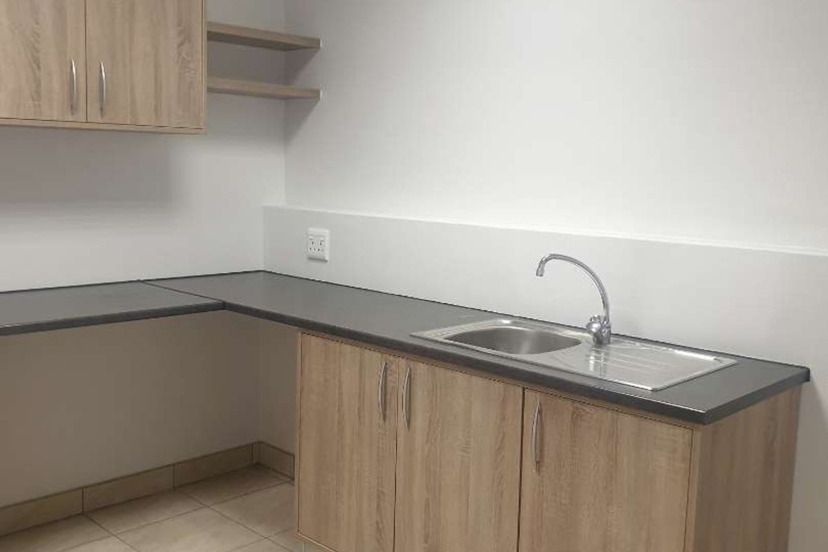 A clean, functional kitchenette area featuring light wood-tone cabinetry, dark laminate countertops, a stainless steel sink, and neutral tiled flooring.