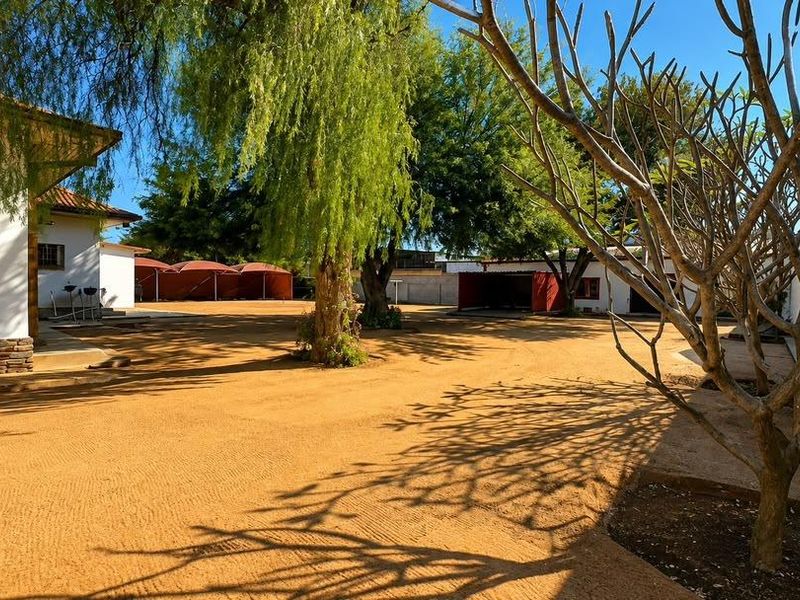 Spacious rear yard area featuring a large open sandy space, mature trees, boundary walls, and covered carport structures.