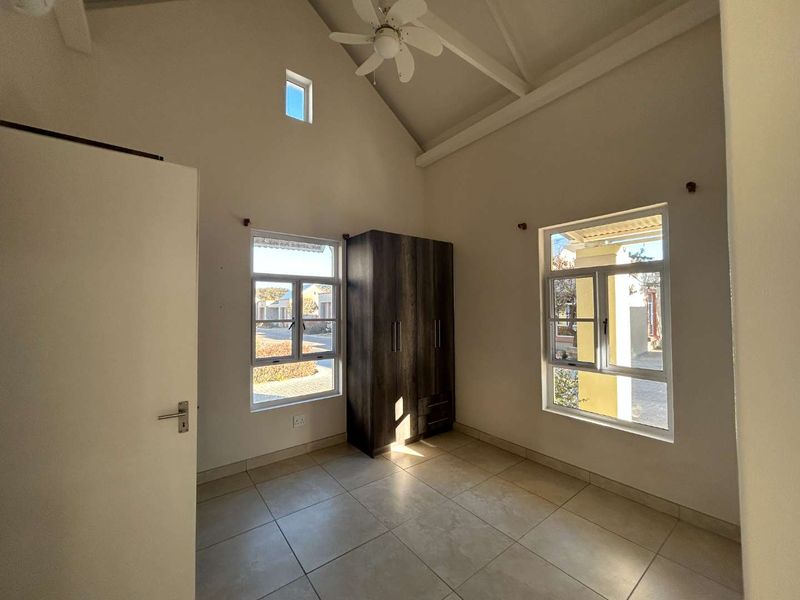 A bright, spacious bedroom featuring a high vaulted ceiling, ceiling fan, tiled flooring, and a built-in wardrobe with large windows providing natural light.