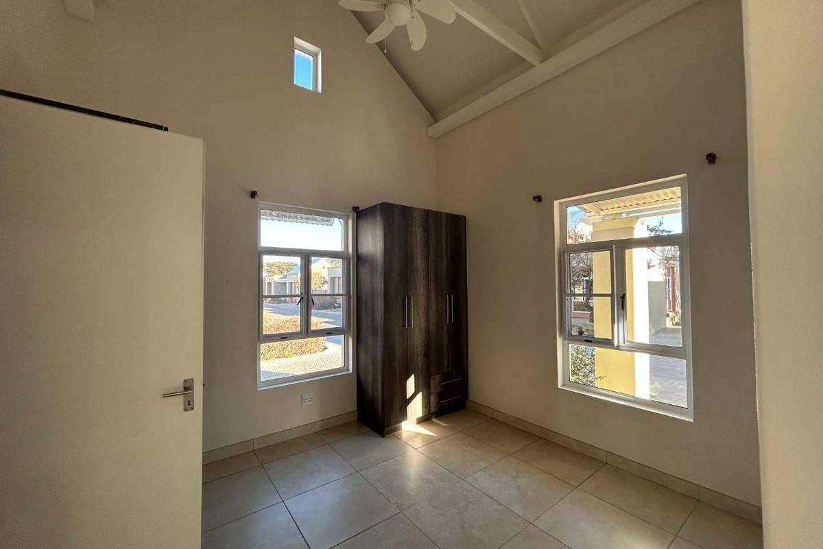A bright, spacious bedroom featuring a high vaulted ceiling, ceiling fan, tiled flooring, and a built-in wardrobe with large windows providing natural light.