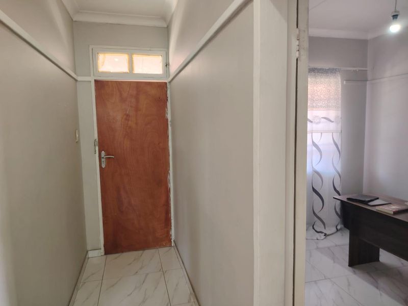 A functional hallway featuring tiled flooring, a wooden entrance door with a transom window, and neutral wall finishes leading into an adjacent room.