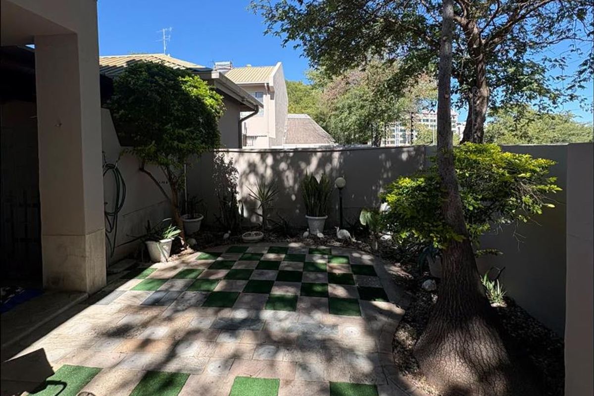 A private rear courtyard featuring a checkerboard-patterned paved patio, mature trees, and high boundary walls for added privacy.