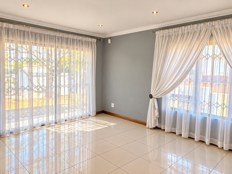 A bright and spacious living area featuring neutral tiled flooring, recessed downlights, and large windows with security gates, offering a clean and modern aesthetic.