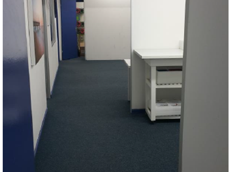 A functional industrial hallway featuring carpeted flooring, partition walls, and overhead fluorescent lighting, providing access to adjacent office or utility spaces.