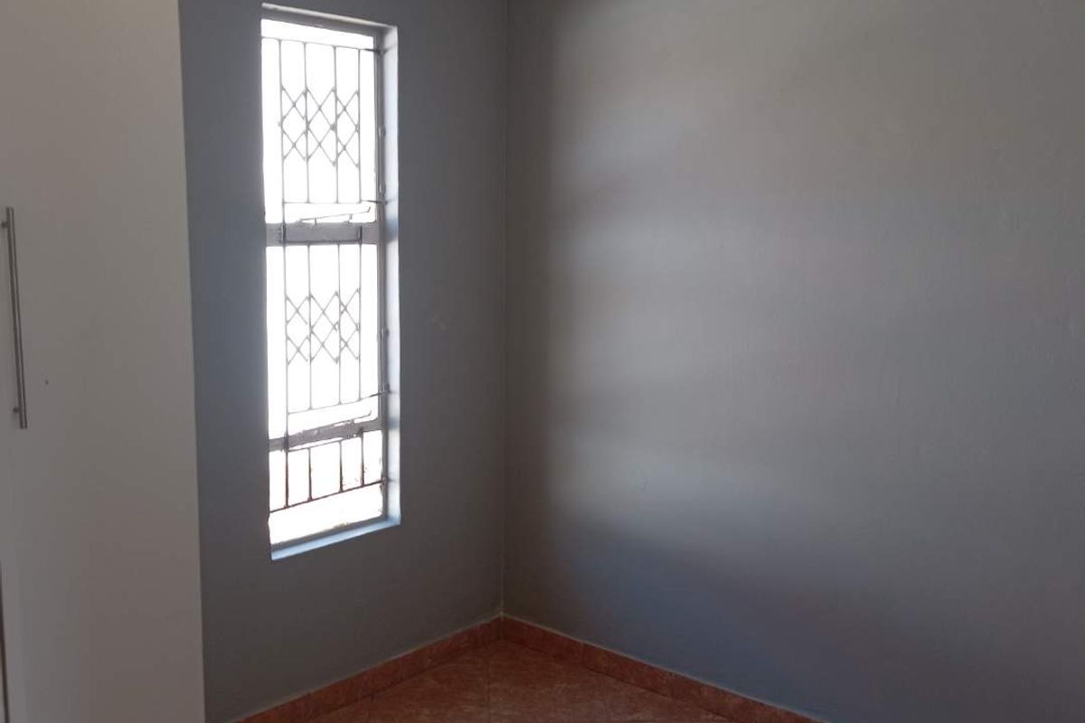 A simple bedroom featuring tiled flooring, grey painted walls, and a window fitted with security burglar bars.
