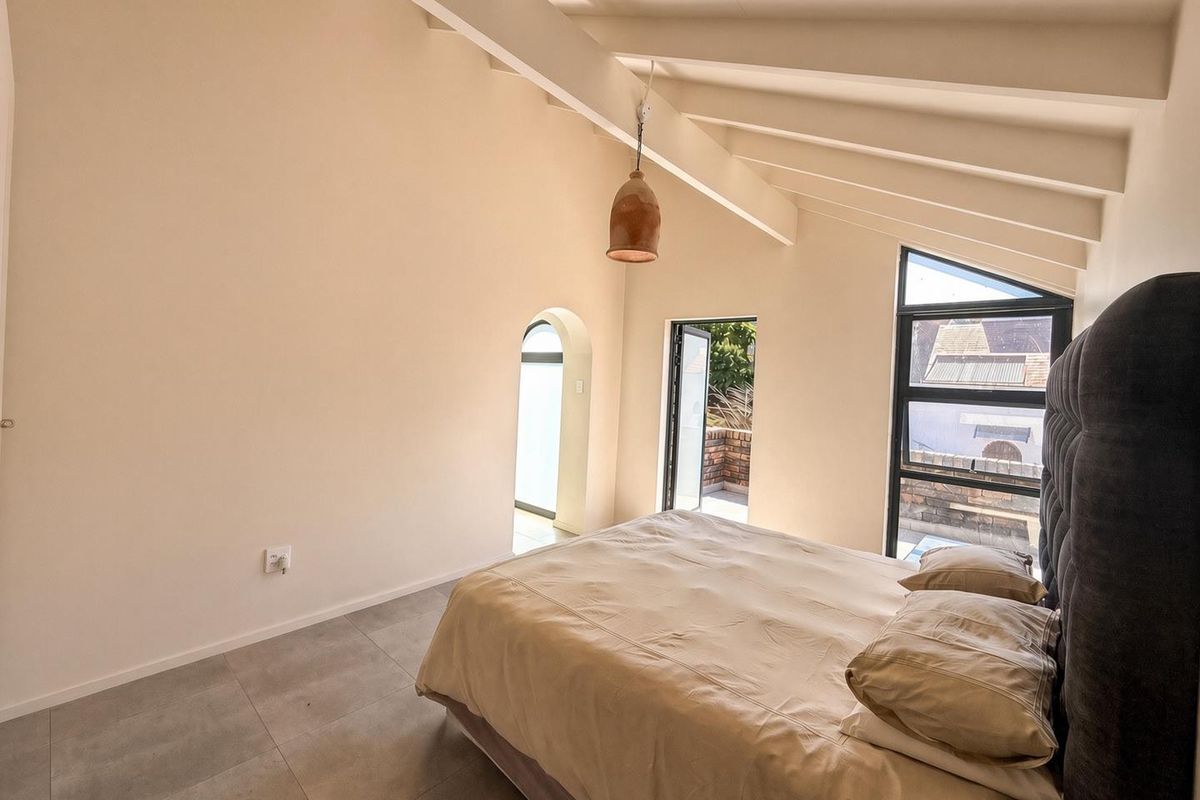 A bright bedroom featuring high ceilings with exposed rafters, tiled flooring, and large windows providing natural light and access to an outdoor area.
