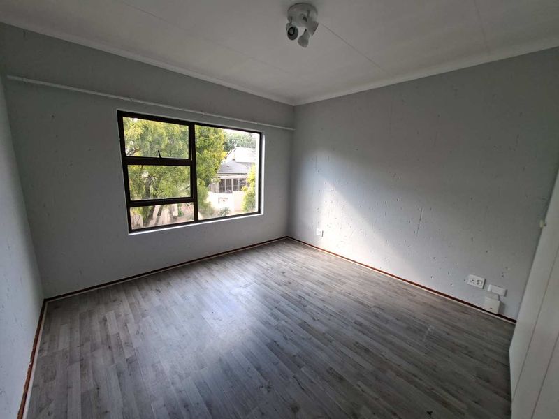 An empty bedroom featuring grey laminate flooring, neutral walls, and a large window providing natural light.