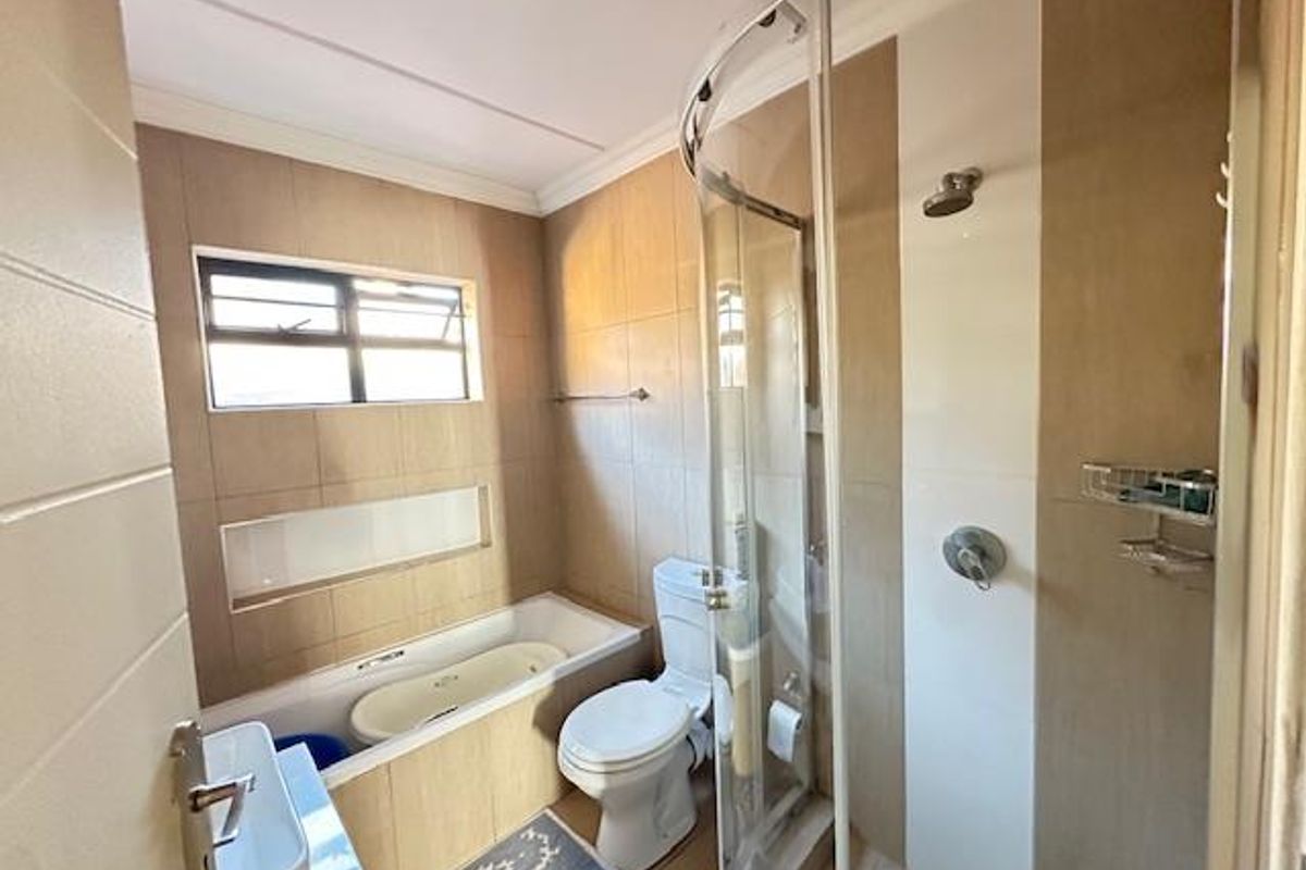 A functional bathroom featuring a glass-enclosed shower, a built-in bathtub, a toilet, and neutral wall tiling with a window for natural light.