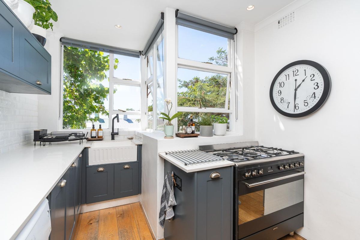 A bright, modern kitchen featuring dark shaker cabinetry, white quartz countertops, a farmhouse sink, and a professional-style gas range with timber flooring.