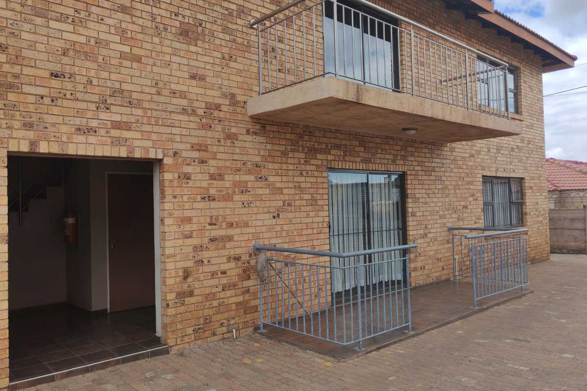 Exterior view of a face brick apartment building featuring a ground floor patio and an upper-level balcony with metal railings.