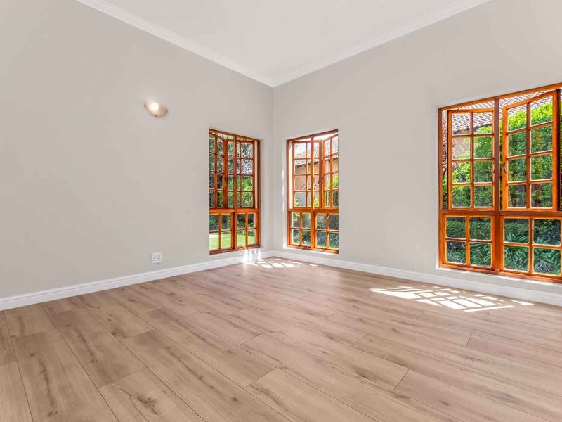 A bright, empty living room featuring modern laminate flooring, neutral walls, and large wooden casement windows overlooking the garden.