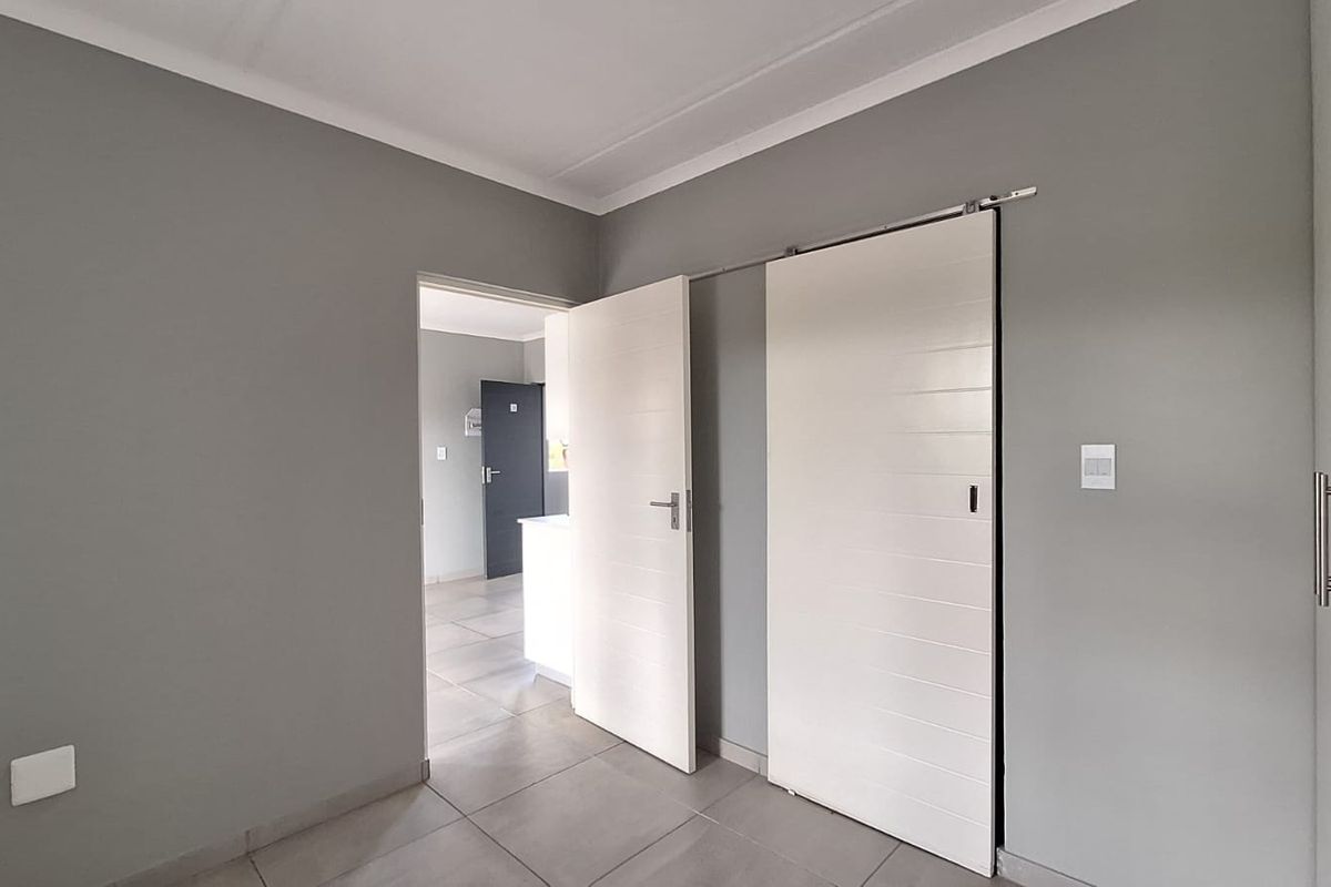 A clean, modern bedroom featuring neutral grey walls, tiled flooring, a sliding door, and built-in cupboard storage.