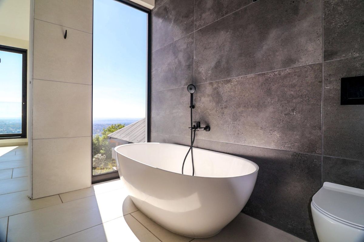 Modern bathroom featuring a freestanding bathtub, wall-mounted shower mixer, large format grey wall tiles, and floor-to-ceiling windows with scenic views.
