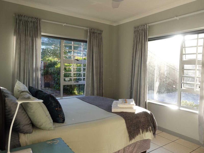 A bright bedroom featuring tiled flooring, large windows with security bars, and a ceiling fan, offering natural light and garden views.