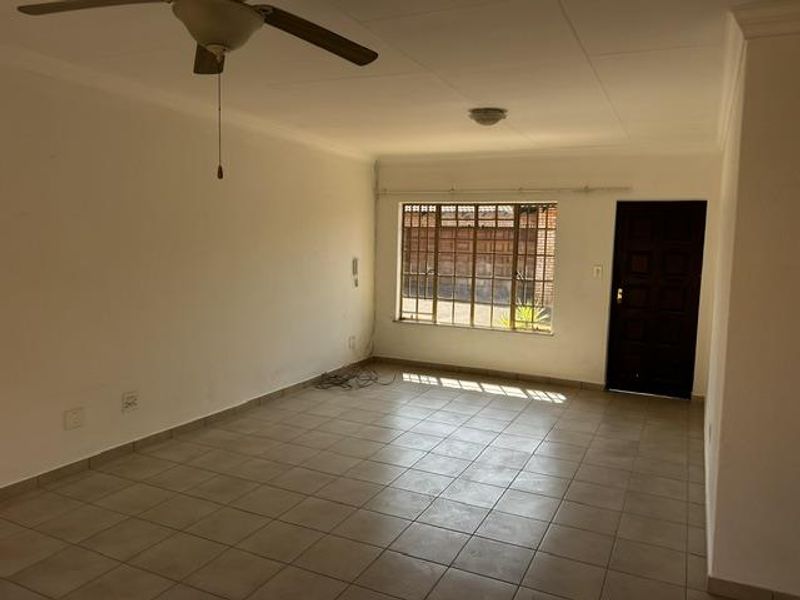 A spacious, empty living area featuring neutral tiled flooring, a ceiling fan, and a secure entrance door with natural light from the window.